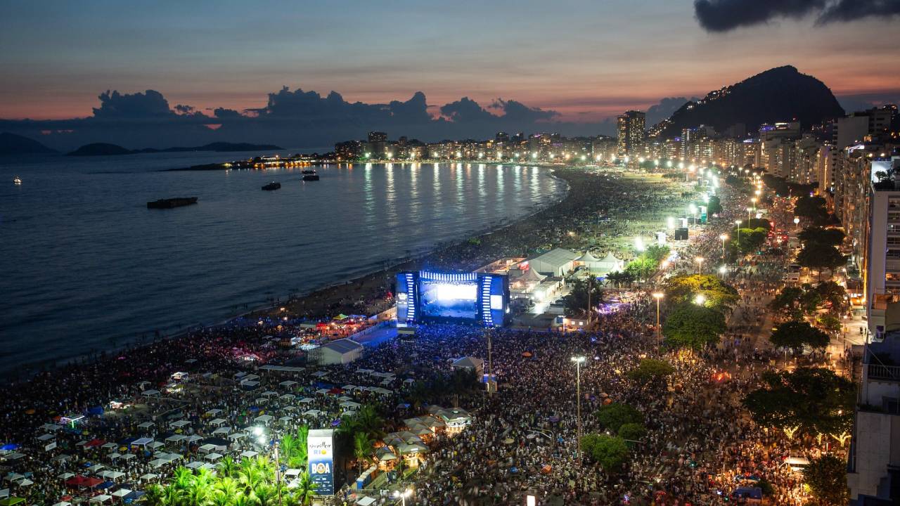 Foto mostra Praia de Copacabana cheia para o Réveillon com um palco instalado
