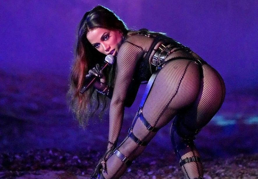 Anitta usa body preto arrastão. Ela está de lado para a câmera, mas seu rosto está de frente