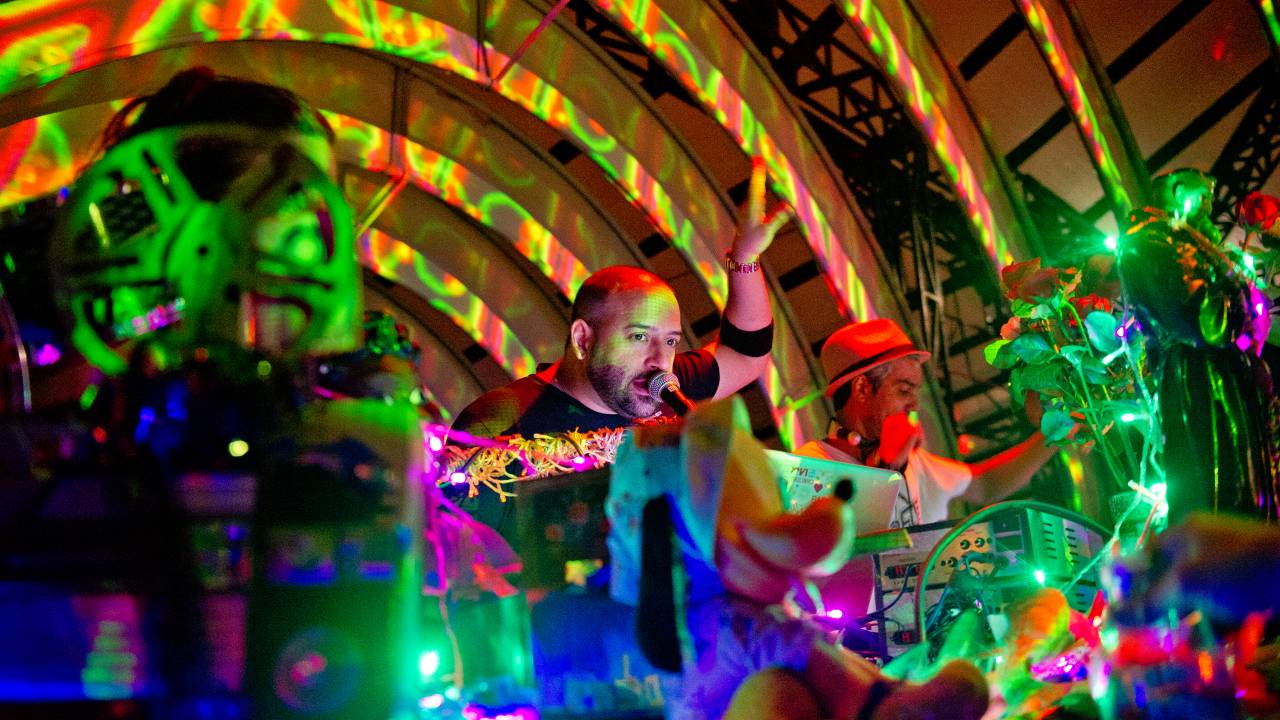A mesa dos DJs com luzes e adornos coloridos. Rodrigo Penna fala ao microfone e Markinhos Mesquita está ao seu lado.