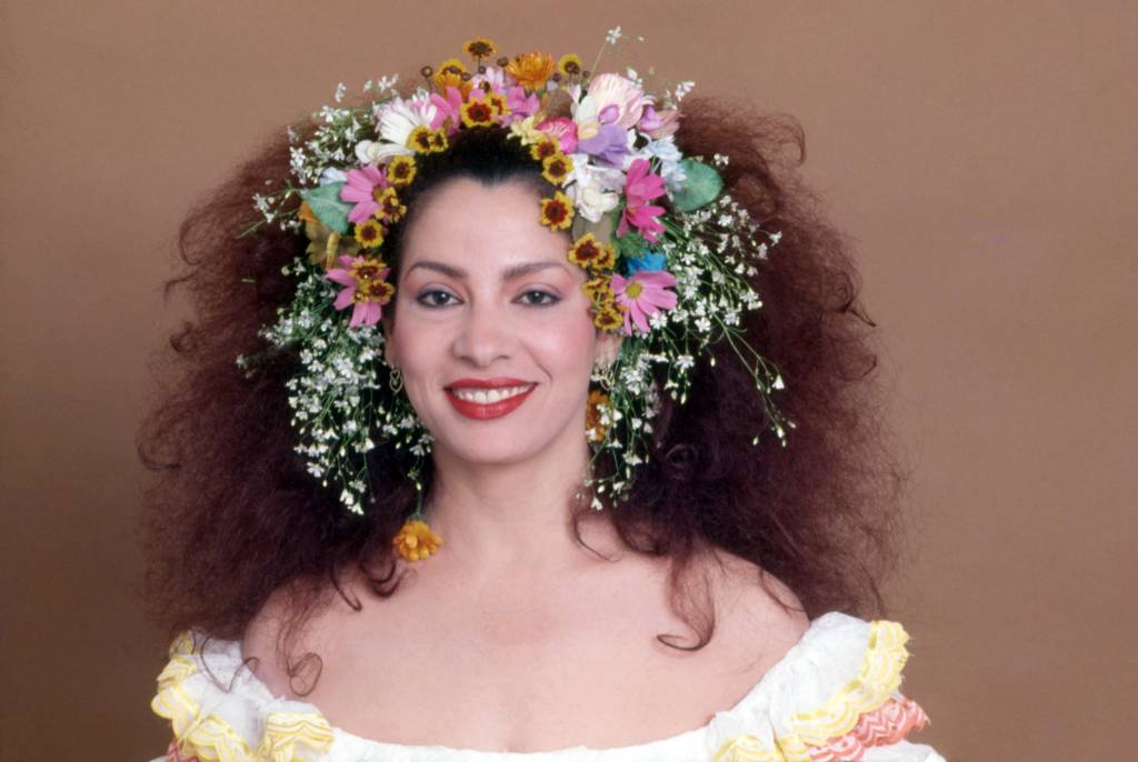 A cantora Clara Nunes de blusa branca com decote ombro a ombro, usando tiara de flores na cabeça. Ela é branca e tem os cabelos castanhos cacheados e volumosos.