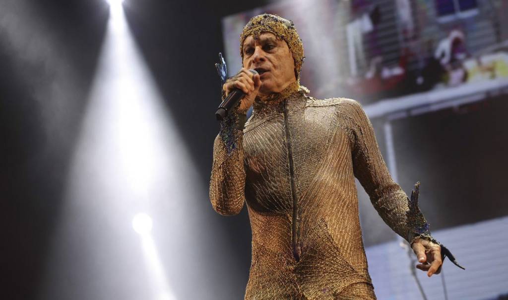 Ney Matogrosso com body suit dourado cantando sob as luzes do palco