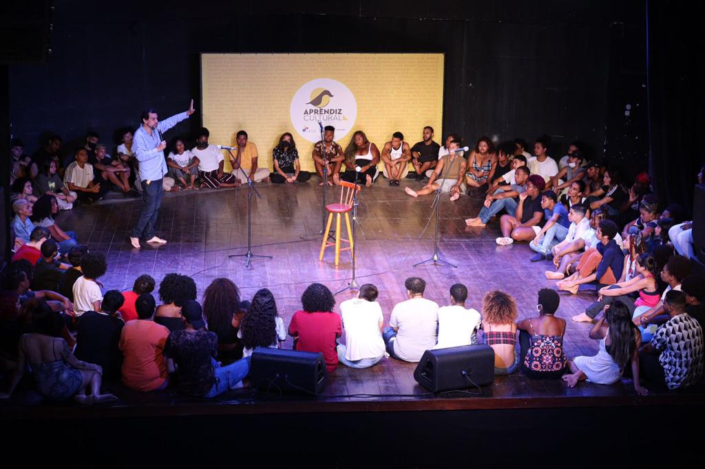 Foto mostra grupo e jovens sentados em círculo ouvndo um homem palestrar no meio