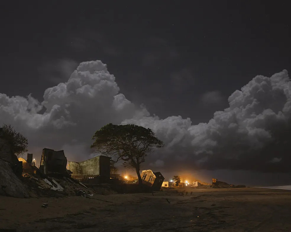 Foto de casas tortas à beira-mar em Atafona, afetadas pelo avanço do mar. Há uma silhueta de uma árvore, uma grande nuvem no céu e luzes ao fundo.