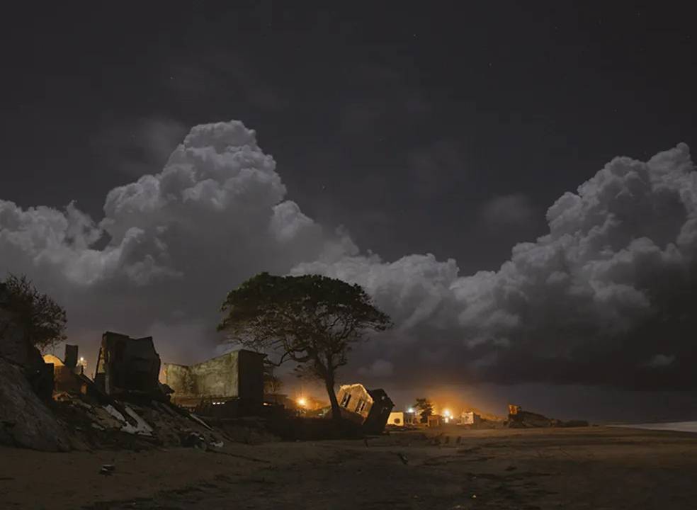 Foto de casas tortas à beira-mar em Atafona, afetadas pelo avanço do mar. Há uma silhueta de uma árvore, uma grande nuvem no céu e luzes ao fundo.