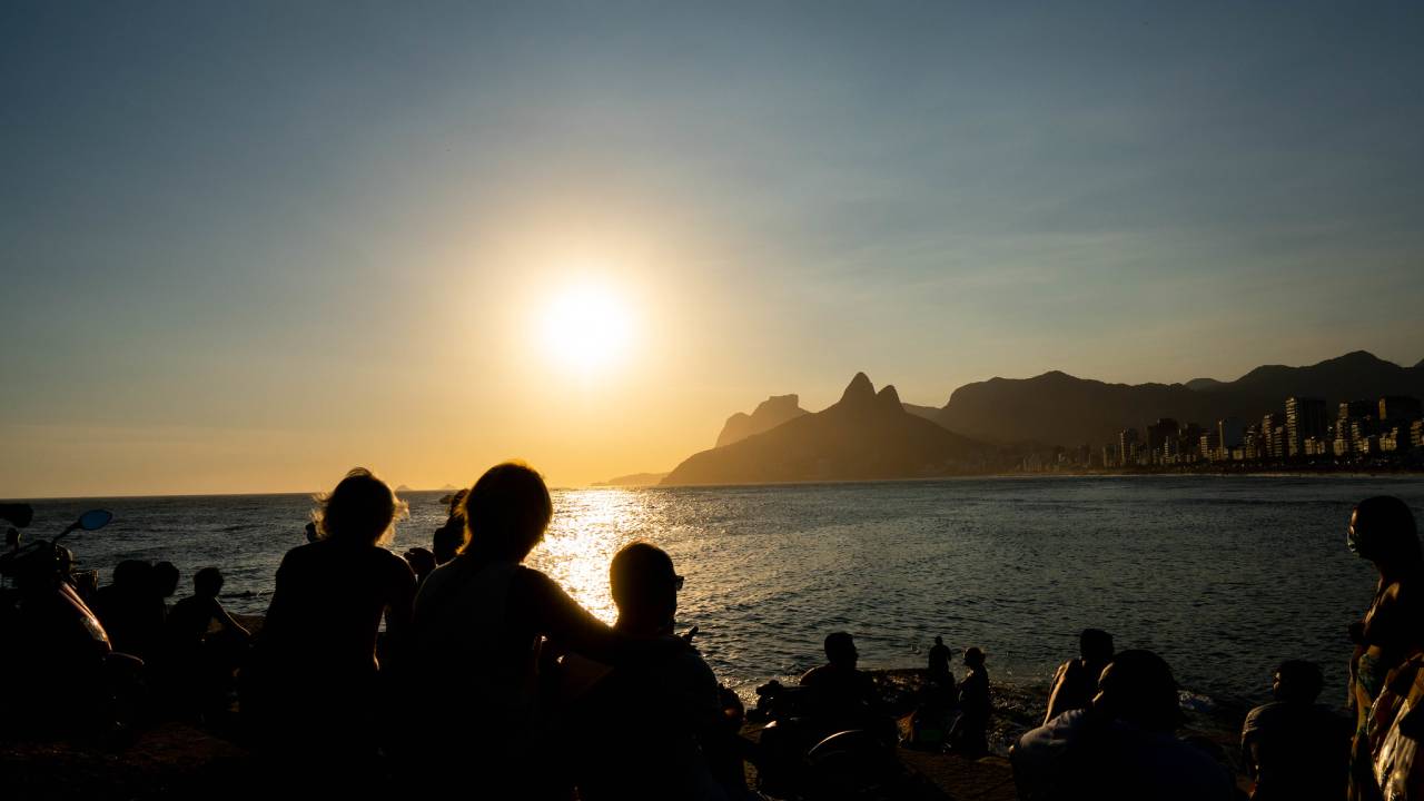 Foto mostra foto de pessoas assistindo ao pôr do sol no Arpoador, olhando em direção ao mar e ao Morro Dois Irmãos