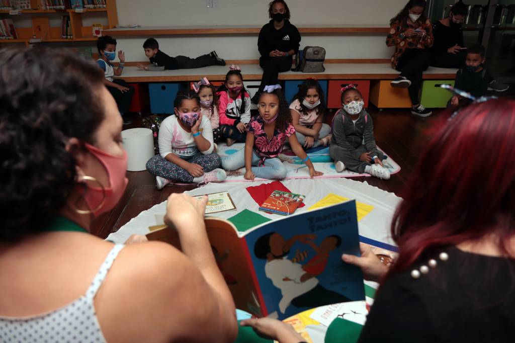 Foto mostra crianças em sessão de contação de histórias. Duas mulheres estão sentadas de frente para elas e segurando um livro aberto
