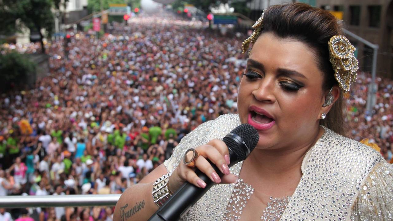 Preta Gil usa roupa bege toda coberta por cristais e arranjo em formato de folhas nas lateiras da cabeça, também beges e com cristais, e canta no trio elétrico à frente de uma multidão que segue o Bloco da Preta