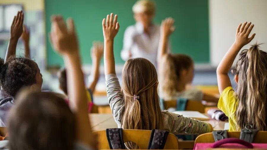 Foto mostra crianças na sala de aula com a mão levantada