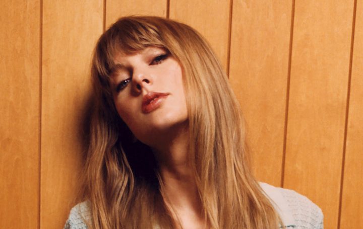 Taylor Swift com os cabelos soltos, de blusa cinza e encostada em uma parede marrom-claro