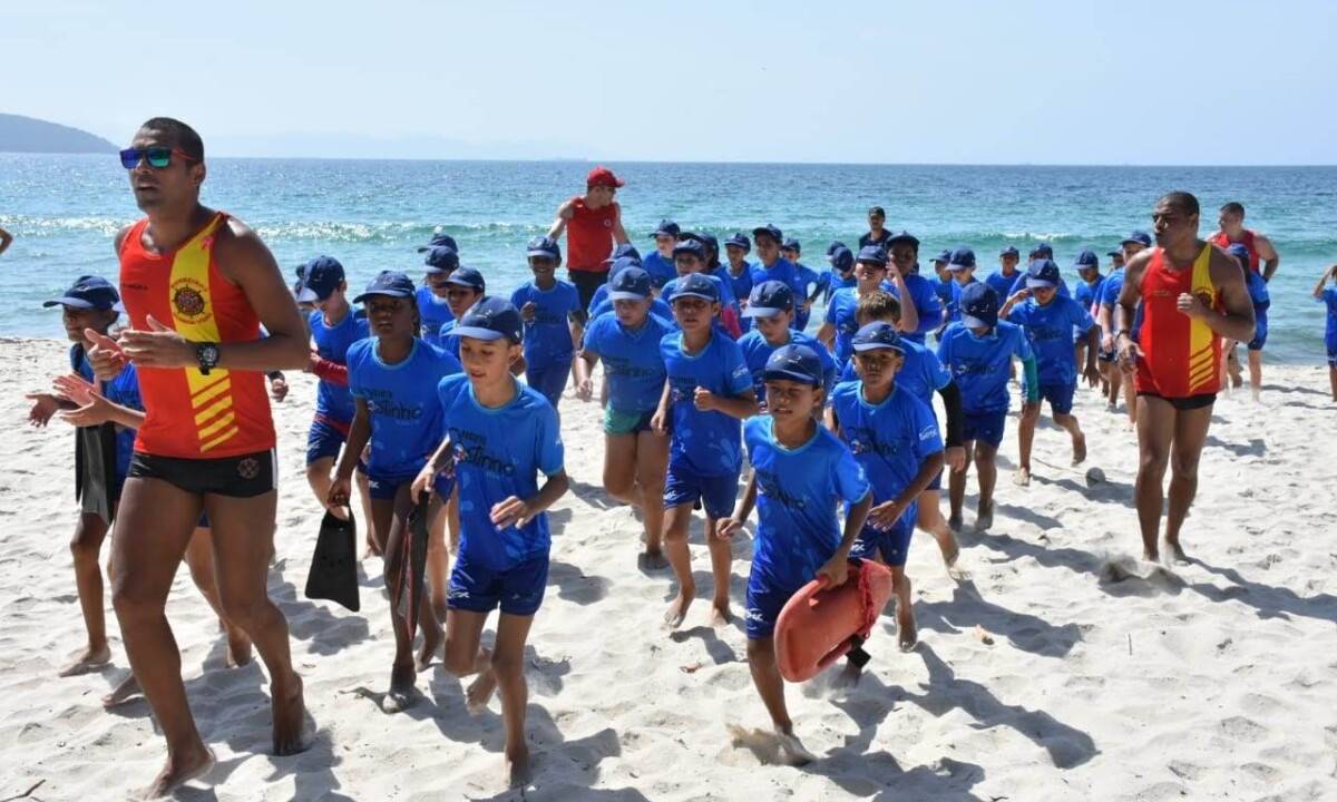 Foto mostra crianças correndo na areia da praia acompanhadas de instrutores do corpo de bombeiros