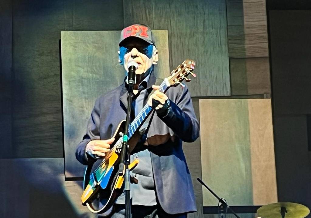 Chico Buarque usa boné com a inscrição CPX. Ele veste calça, camisa e paletó azuis e toca violão elétrico vazado.
