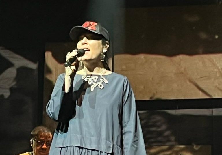 Monica Salmaso está segurando o microfone e cantando. Ela usa vestido azul-acinzentado e boné preto com a inscrição CPX em vermelho.