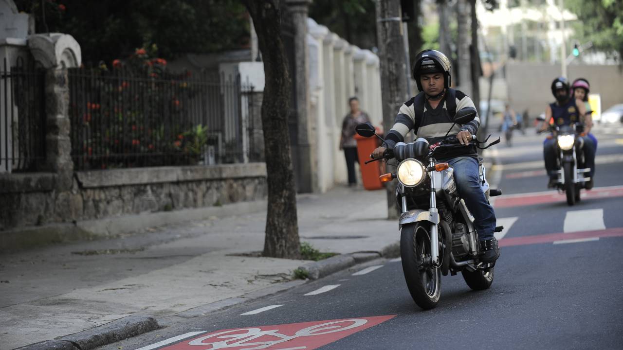 Foto mostra um homem em uma moto à frente e, atrás, uma moto com motorista e passageiro.