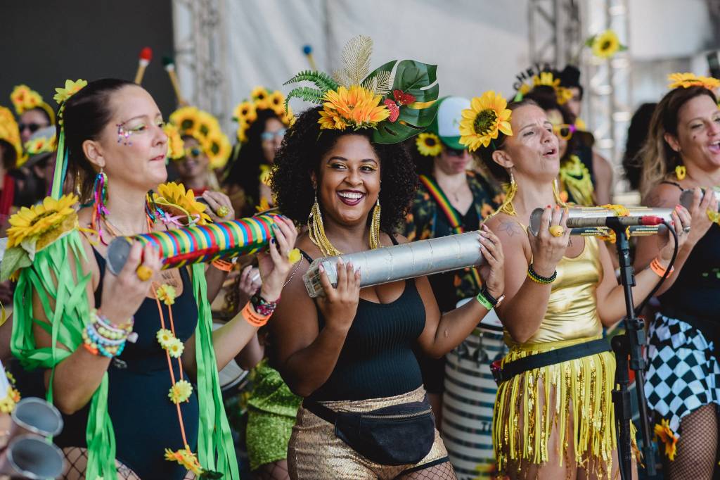 Mulheres instrumentistas do bloco Que Pena Amor com girassois na cabeça e tocando chocalho.