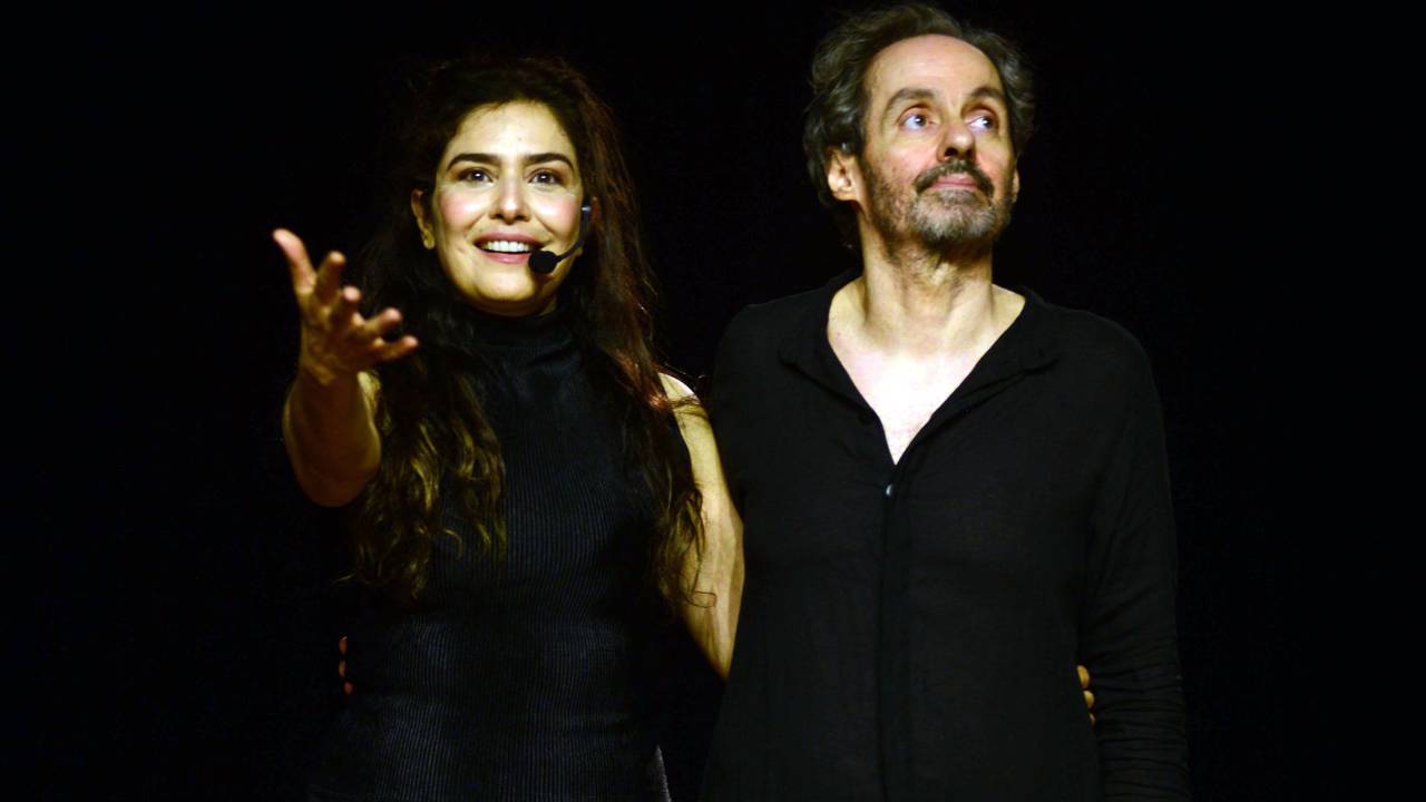 Letícia Sabatella e Daniel Dantas vestem roupas pretas sobre fundo preto. Ela sorri, usa microfone e aponta com a mão direita para a frente.