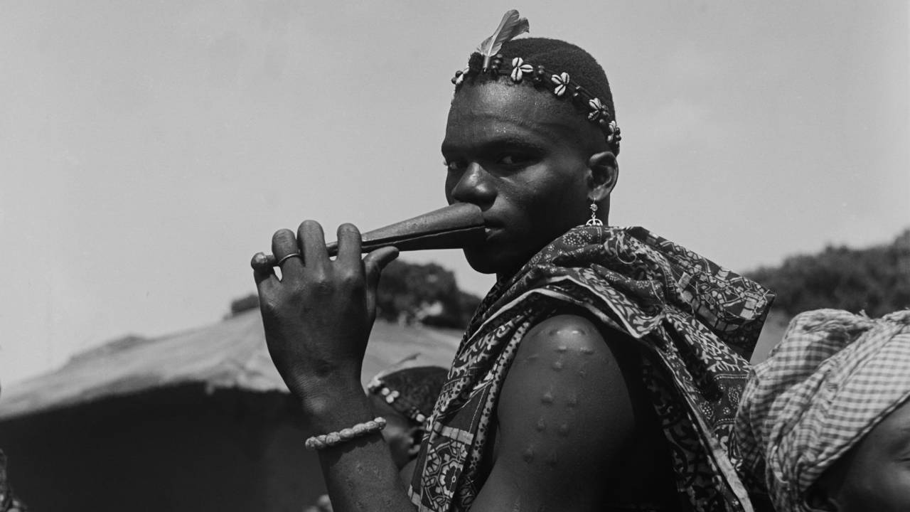 Foto em preto e branco. Homem negro do Benim com cabelo curto e adereço na cabeça. pulseira, brinco, tecido estampado sobre o ombro. Seu corpo está de lado e ele olha para a câmera, assoprando um apito ou instrumento musical.