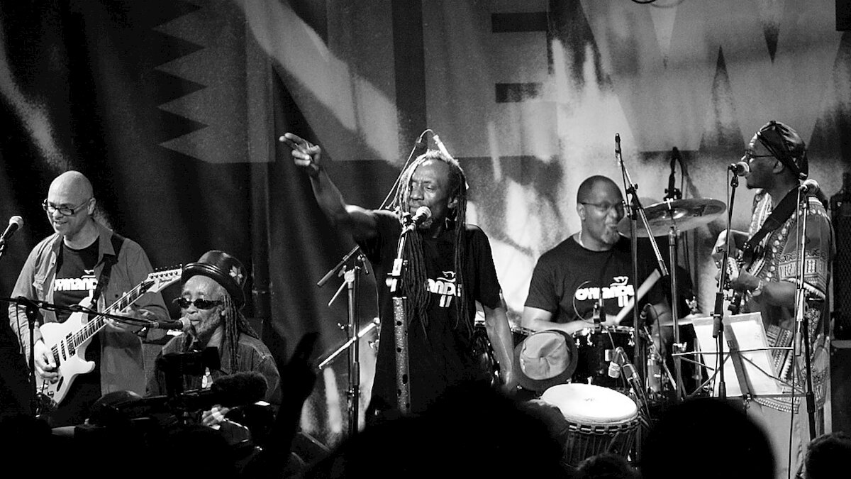Foto em preto e branco de cinco integrantes da banda Cymande em ação, no palco