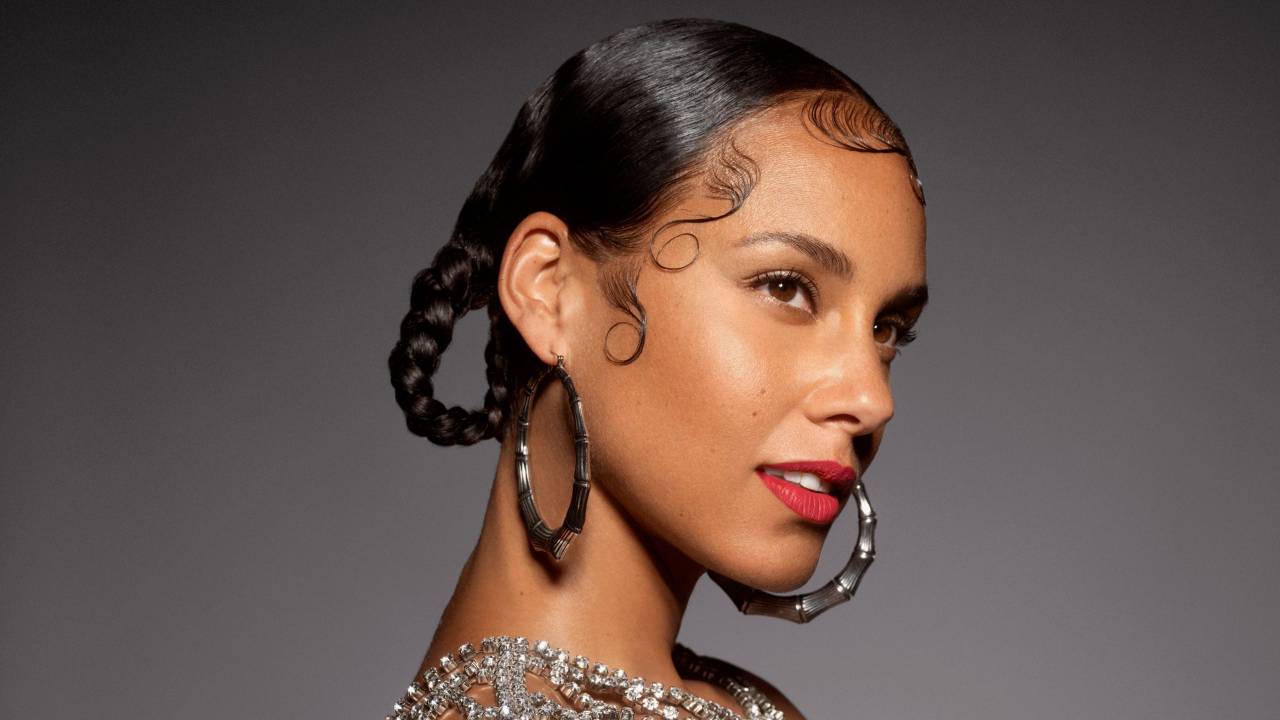 Alicia Keys sobre fundo cinza, com cabelo preso e finalizado com duas tranças presas, argolas grandes prateadas e blusa de cristais.