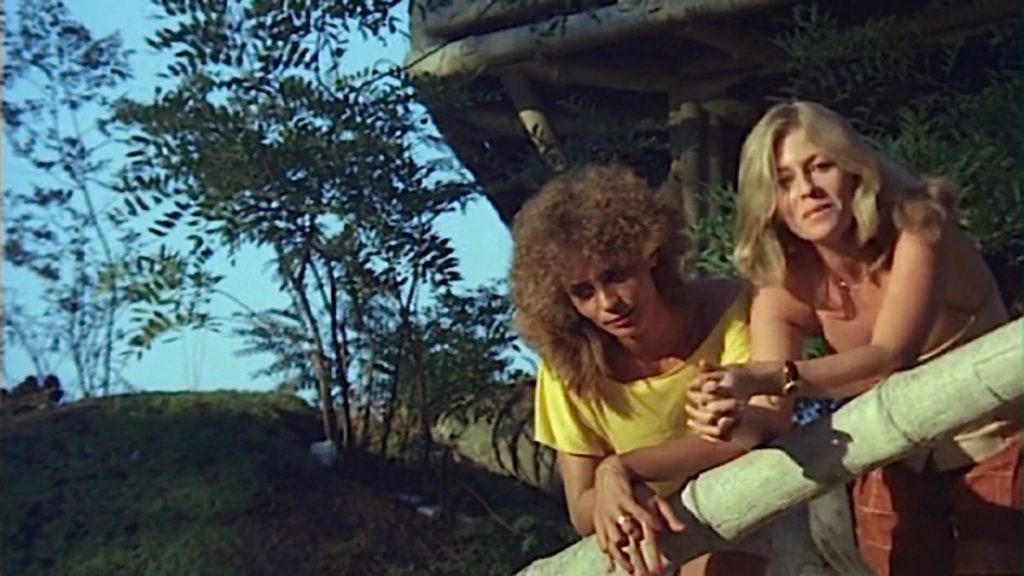 Duas mulheres estão apoiadas em uma varanda. À esquerda, a atriz Wilma Dias, que tem o cabelo loiro escuro e cacheado, ela veste blusa amarela e olha para baixo. À direita, a atriz Monique Laffond, que é loira de cabelos lisos e olha para a frente, esboçando um discreto sorriso.