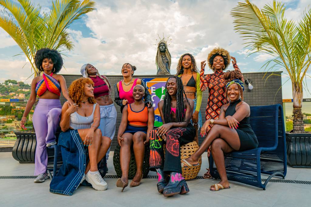 Nove mulheres negras juntas e sorrindo, algumas em pé, algumas sentadas em cadeiras ou pufes