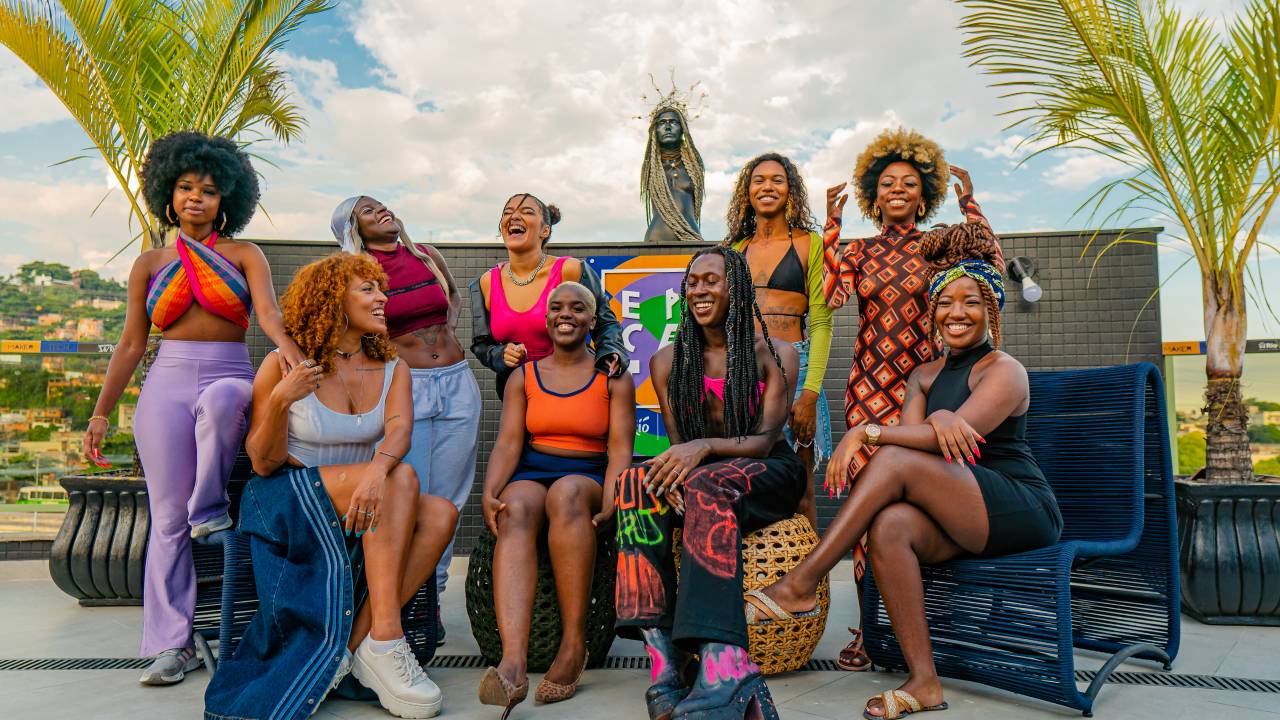 Nove mulheres negras juntas e sorrindo, algumas em pé, algumas sentadas em cadeiras ou pufes