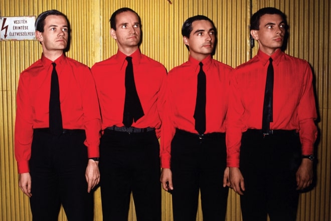 Os quatro integrantes do Kraftwerk de calça e gravata preta e camisa vermelha, encostados em uma parede bege.