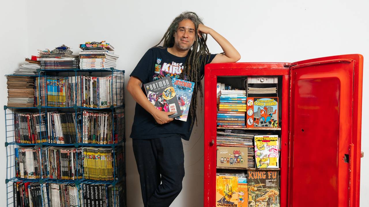 Lencinho é um homem branco de dreadlocks grandes. Ele está entre sua geladeira vermelha, com a porta aberta e cheia de HQs, e uma estante cheia de livros