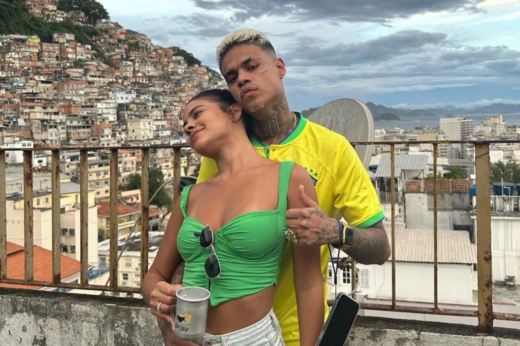 MC Cabelinho usa camisa da seleção brasileira. Ele abraça Bella Campos por trás, e ela usa top verde e segura uma caneca na mão. Ao fundo, a vista para uma favela.
