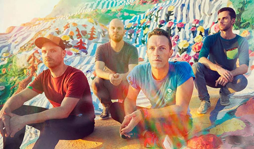 Os quatro integrantes do Coldplay sentados no chão em uma foto com muitas cores e raios de sol sobre os artistas.