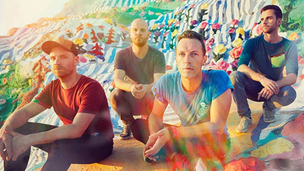 Os quatro integrantes do Coldplay sentados no chão em uma foto com muitas cores e raios de sol sobre os artistas.