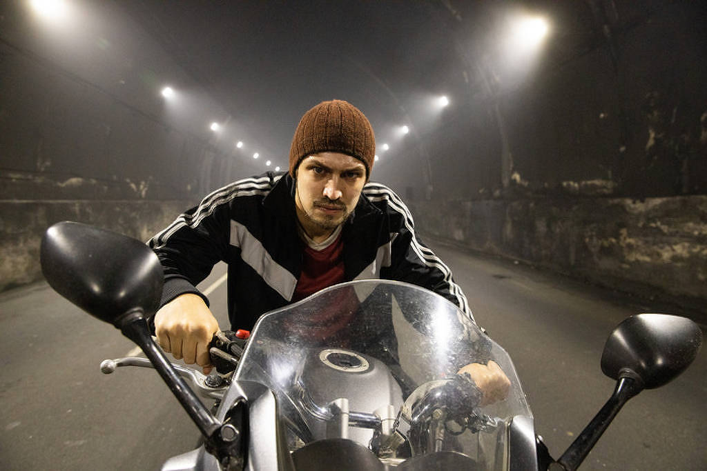 Gabriel Leone caracterizado de Pedro Dom, com gorro, jaqueta e em cima de uma moto dentro de um túnel