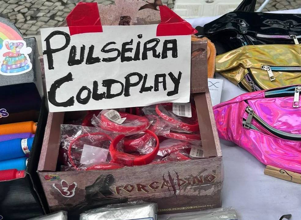 Foto de objetos à venda no chão, entre eles uma caixa com pulseiras e a placa: "Pulseira Coldplay".