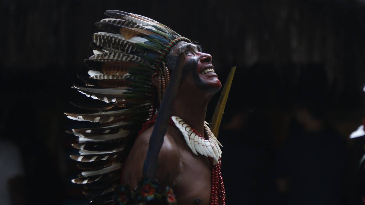 Homem indígena com vistoso cocar está sorrindo e olhando para o alto