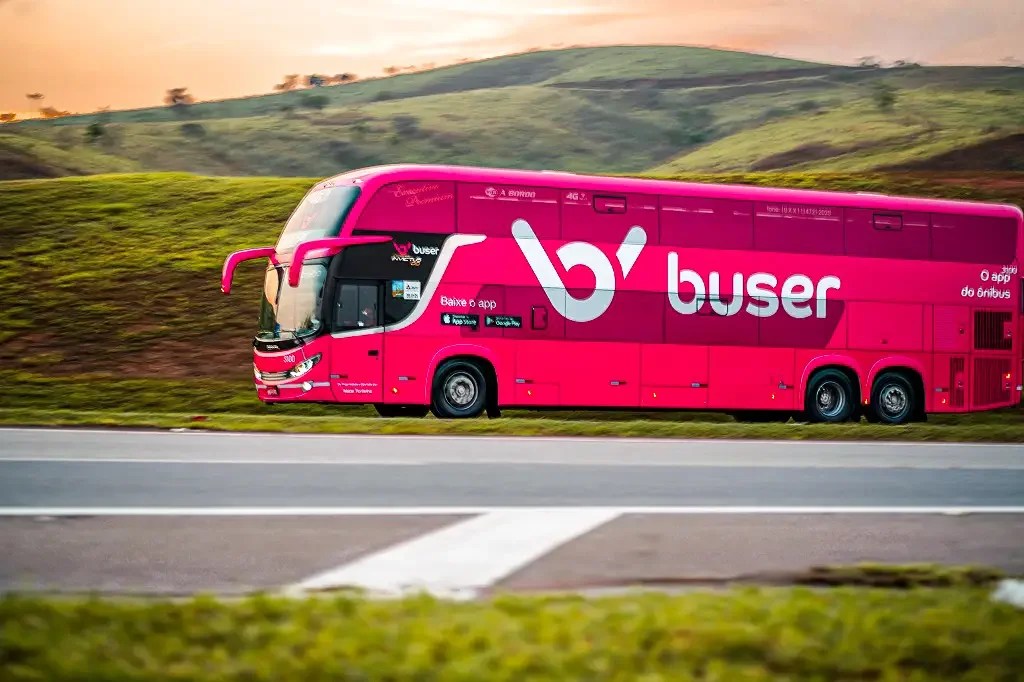 Foto mostra ônibus cor de rosa com o logo da empresa Buser passando por uma estrada cercada por montanhas