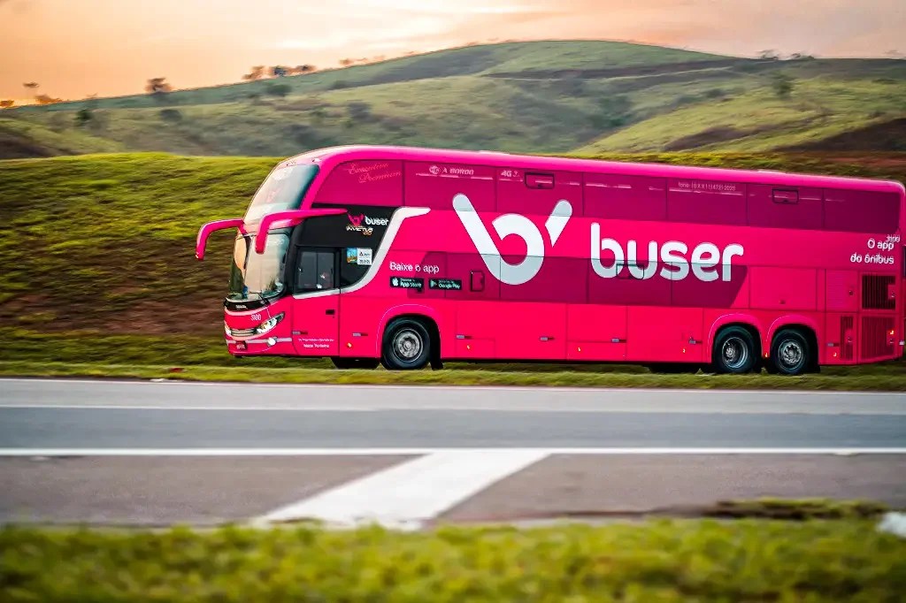 Foto mostra ônibus cor de rosa com o logo da empresa Buser passando por uma estrada cercada por montanhas