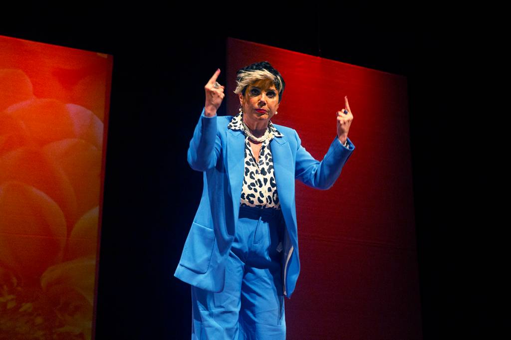 Heloisa Périssé está em cima de um palco com fundo vermelho, de terno azul e com camisa de oncinha discursando.