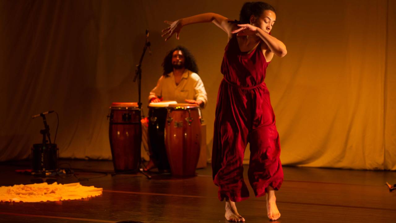 Em primeiro plano, uma dançarina negra pula, usando roupa vinho. Ao fundo, um músico de cabelos compridos toca três atabaques.