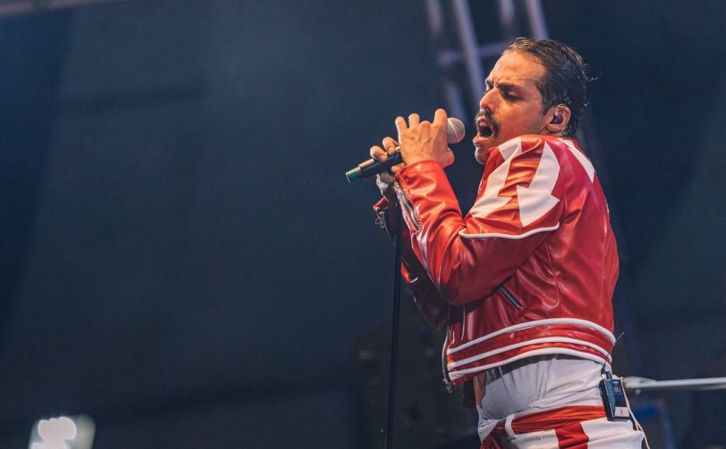 Cover de Freddie Mercury está cantando ao microfone de calça jeans clara e jaqueta vermelha com detalhes brancos