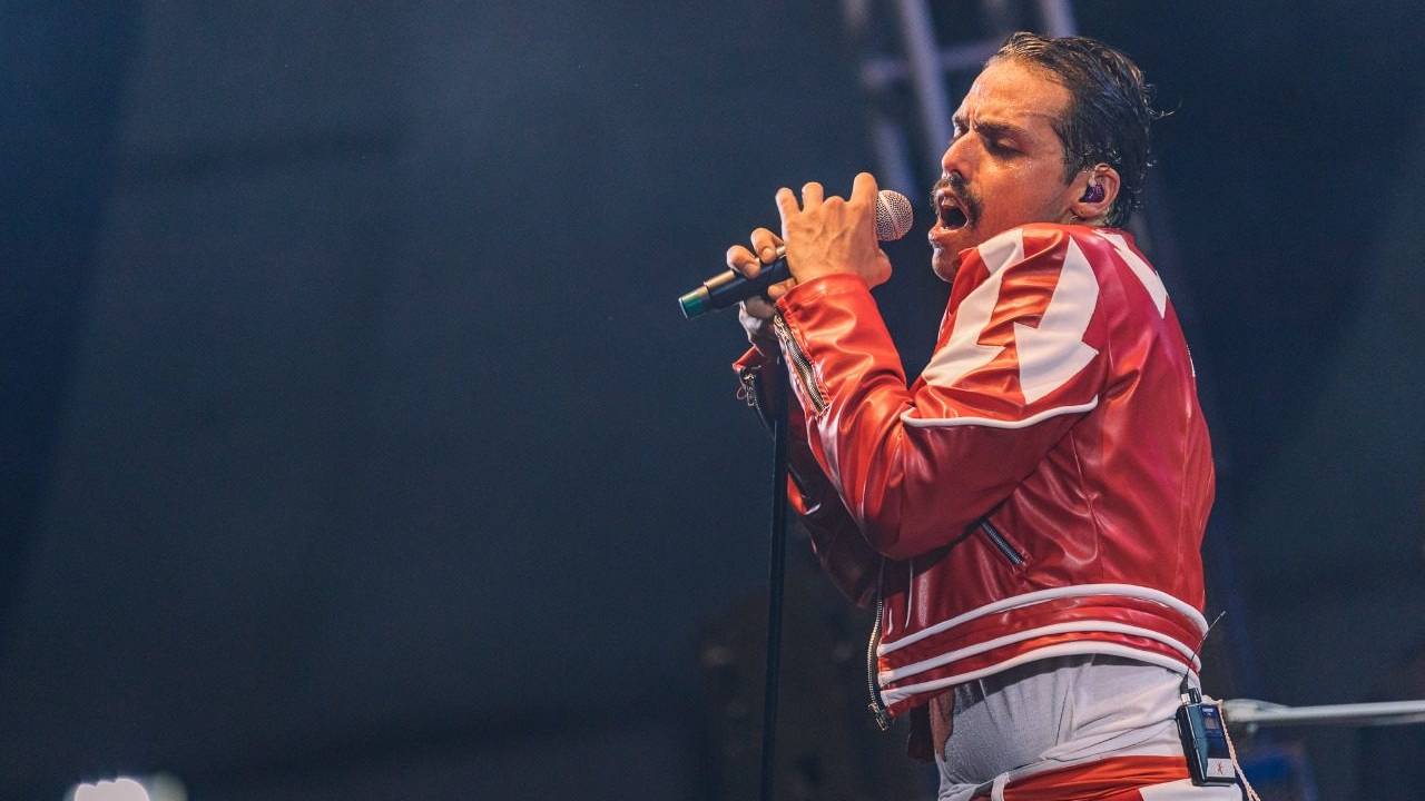 Cover de Freddie Mercury está cantando ao microfone de calça jeans clara e jaqueta vermelha com detalhes brancos