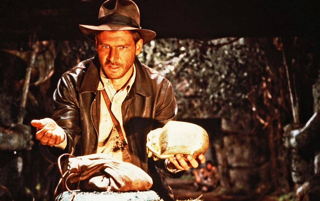 Harrison Ford caracterizado como o arqueólogo Indiana Jones, com roupas em tom de marrom, incluindo chapéu, e segurando um objeto