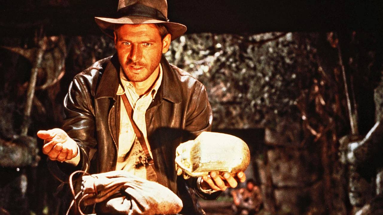 Harrison Ford caracterizado como o arqueólogo Indiana Jones, com roupas em tom de marrom, incluindo chapéu, e segurando um objeto