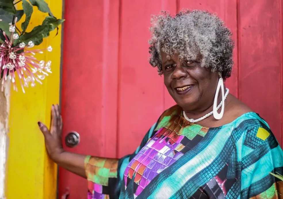 A escritora Conceição Evaristo é uma mulher negra com cabelos grisalhos cacheados. Ela está olhando para a câmera e sorrindo. Ela está usando blusa estampada com tons de azul, usa um brinco branco em formato de gota vazada e está na frente de uma porta vermelha com batente amarelo.