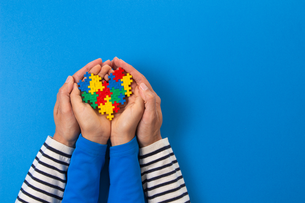 Mãos de adulto e de crianças seguram um coração de brinquedo, reproduzindo a logomarca do autismo.