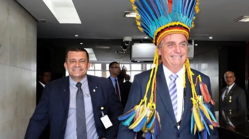 Foto mostra Bolsonaro usando um cocar indígena na cabeça e eu segurança ao lado, ambos usando paletó, camisa social e gravata