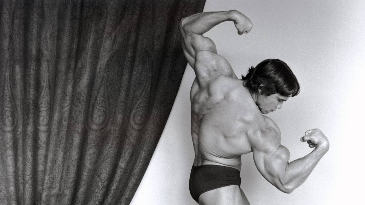 O ator Arnold Schwarzenegger é um homem branco de cabelos curtos. Na foto em preto e branco, ele está de sunga, de lado, fazendo pose com os braços musculosos
