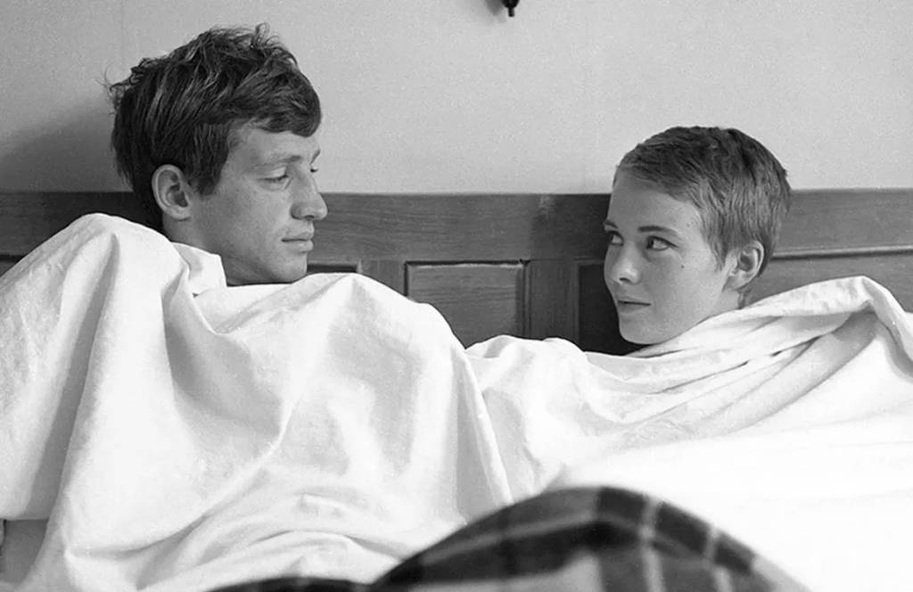 Foto em preto e branco de um casal sentado na cama e coberto com um lençol branco. À esquerda, Jean-Paul Belmondo, branco, de cabelo curto escuro, olha para Jean Seberg, de cabelos curtos e claros