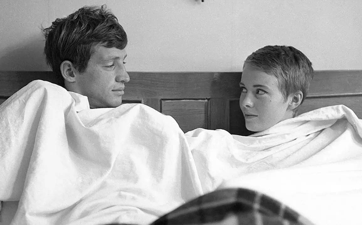 Foto em preto e branco de um casal sentado na cama e coberto com um lençol branco. À esquerda, Jean-Paul Belmondo, branco, de cabelo curto escuro, olha para Jean Seberg, de cabelos curtos e claros