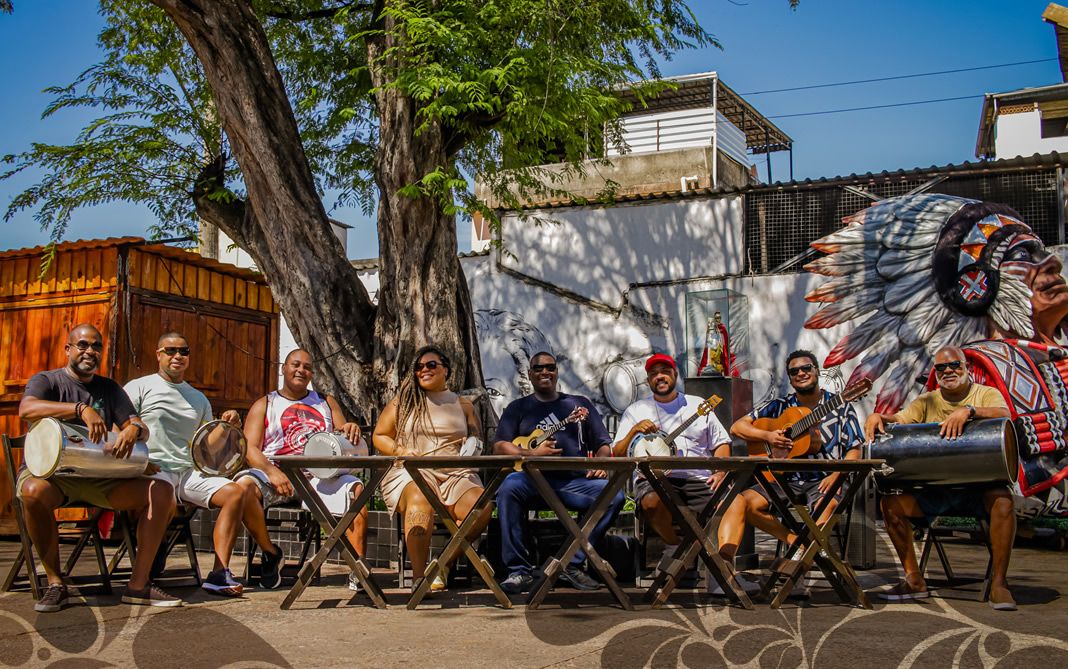 Foto mostra sambistas debaixo de árvore tocando samba