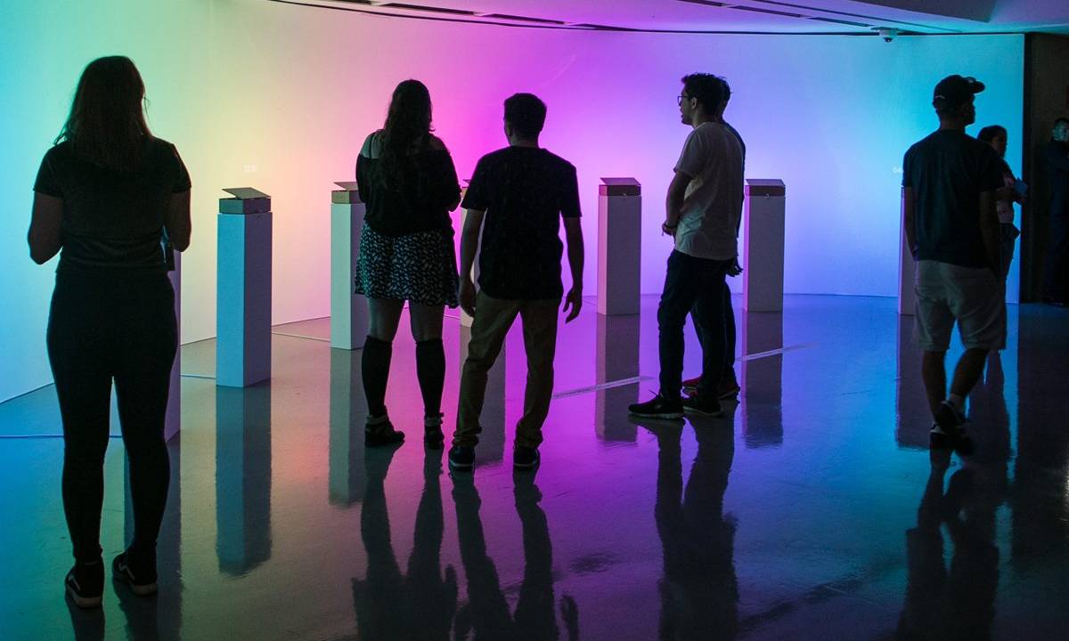 Silhuetas de pessoas em uma sala iluminada com luzes coloridas e barras da instalação artística