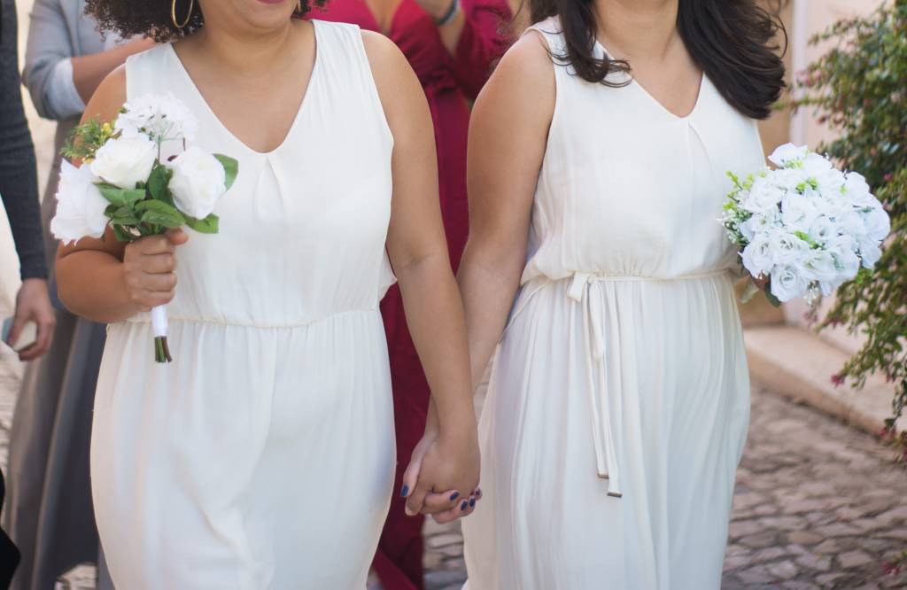Foto mostra uma mulher negra e uma mulher branca de mãos dadas casando, cada uma usando vestido branco e buquê de rosas brancas